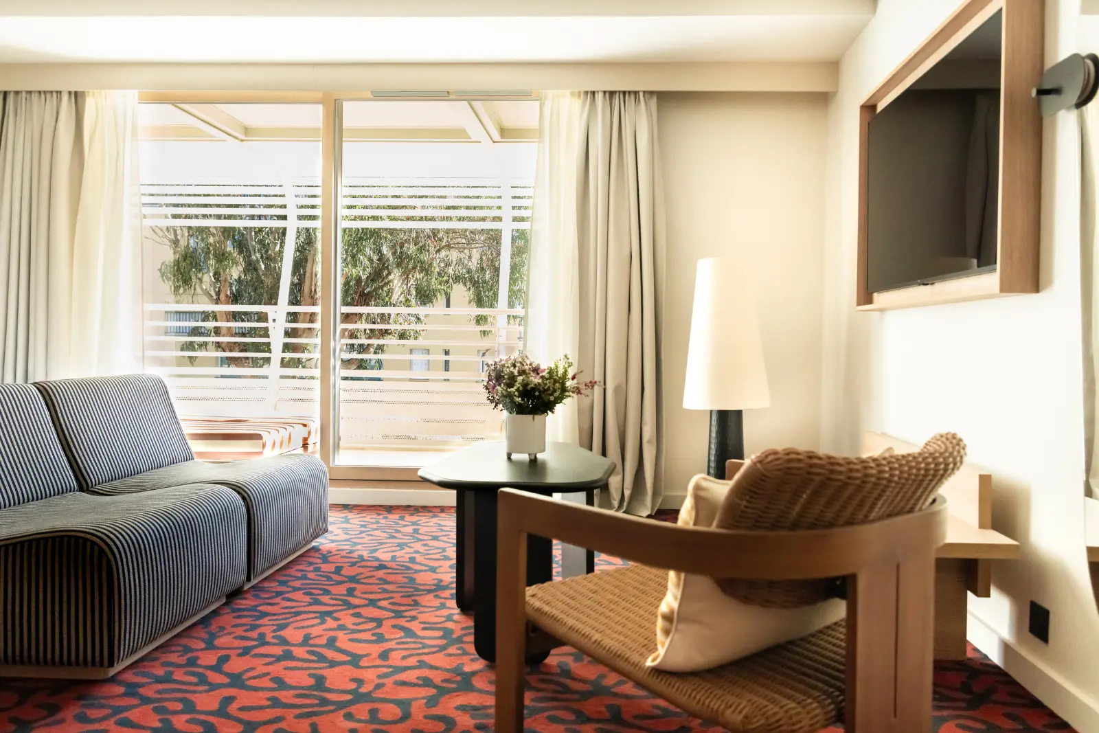 Hotel lounge area with a wooden and rattan armchair, a black side table, a floor lamp and a bay window opening onto a balcony