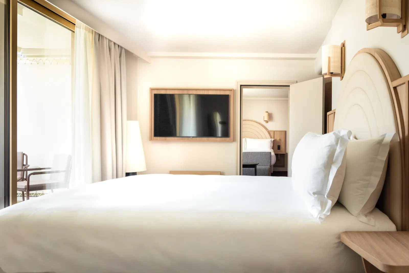 A bright hotel room with a double bed made up with white sheets, a TV in a wooden frame and access to the balcony