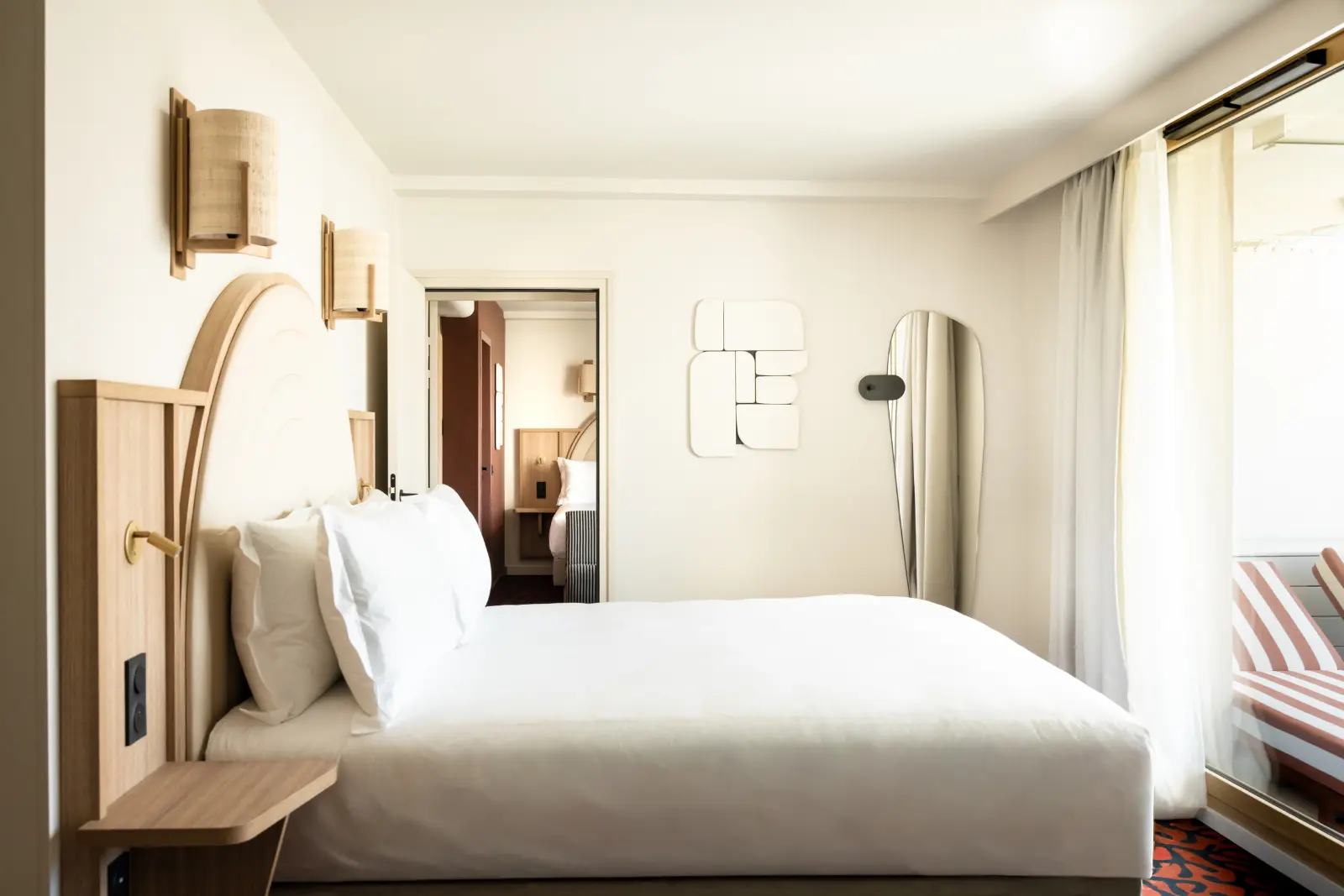 A stylish hotel room with a double bed dressed in white linen, a light wood headboard with arches, rattan wall lights and an oval wall mirror