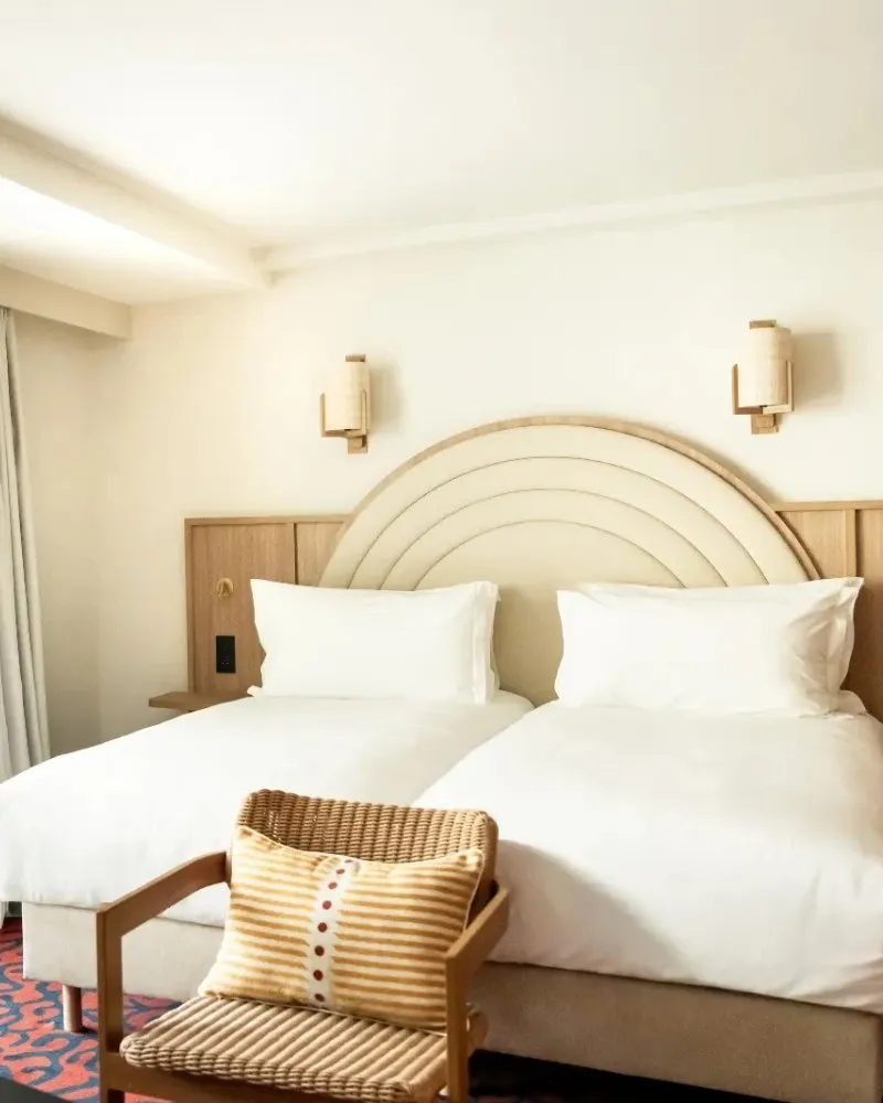 Chambre twin d'hôtel avec deux lits simples, tête de lit aux arches concentriques en bois clair, appliques en rotin et fauteuil en bois avec coussin rayé