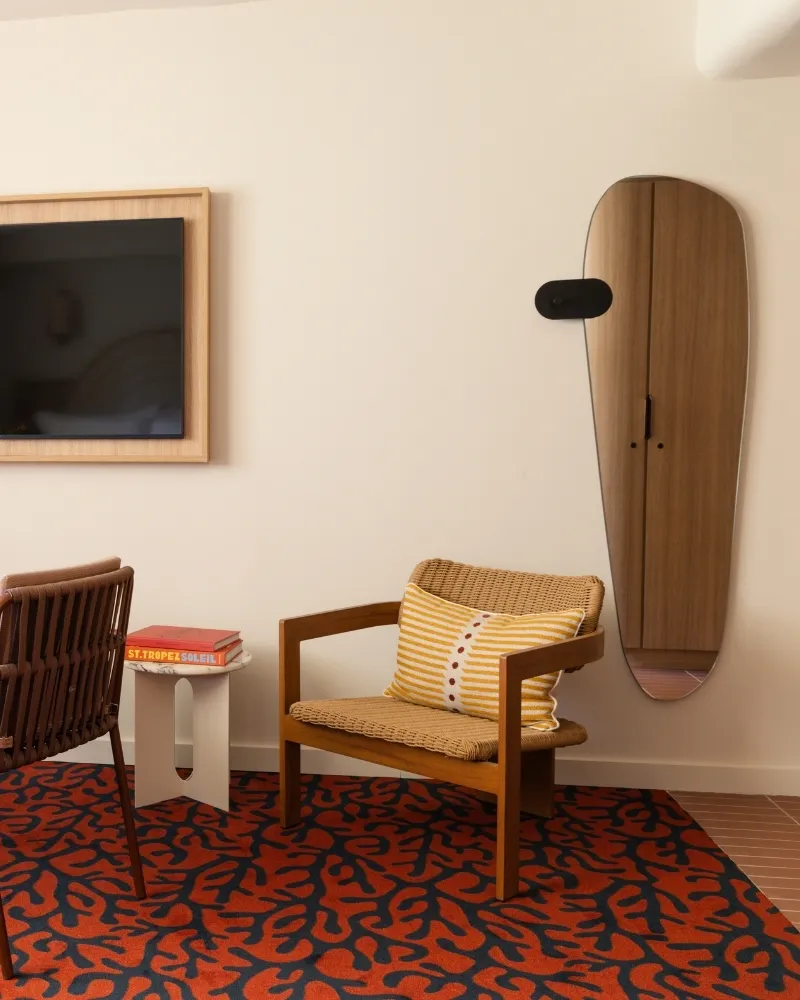 Chambre d'hôtel design avec télévision encadrée en bois, miroir ovale, Moquette rouge aux motifs organiques et fauteuil en osier