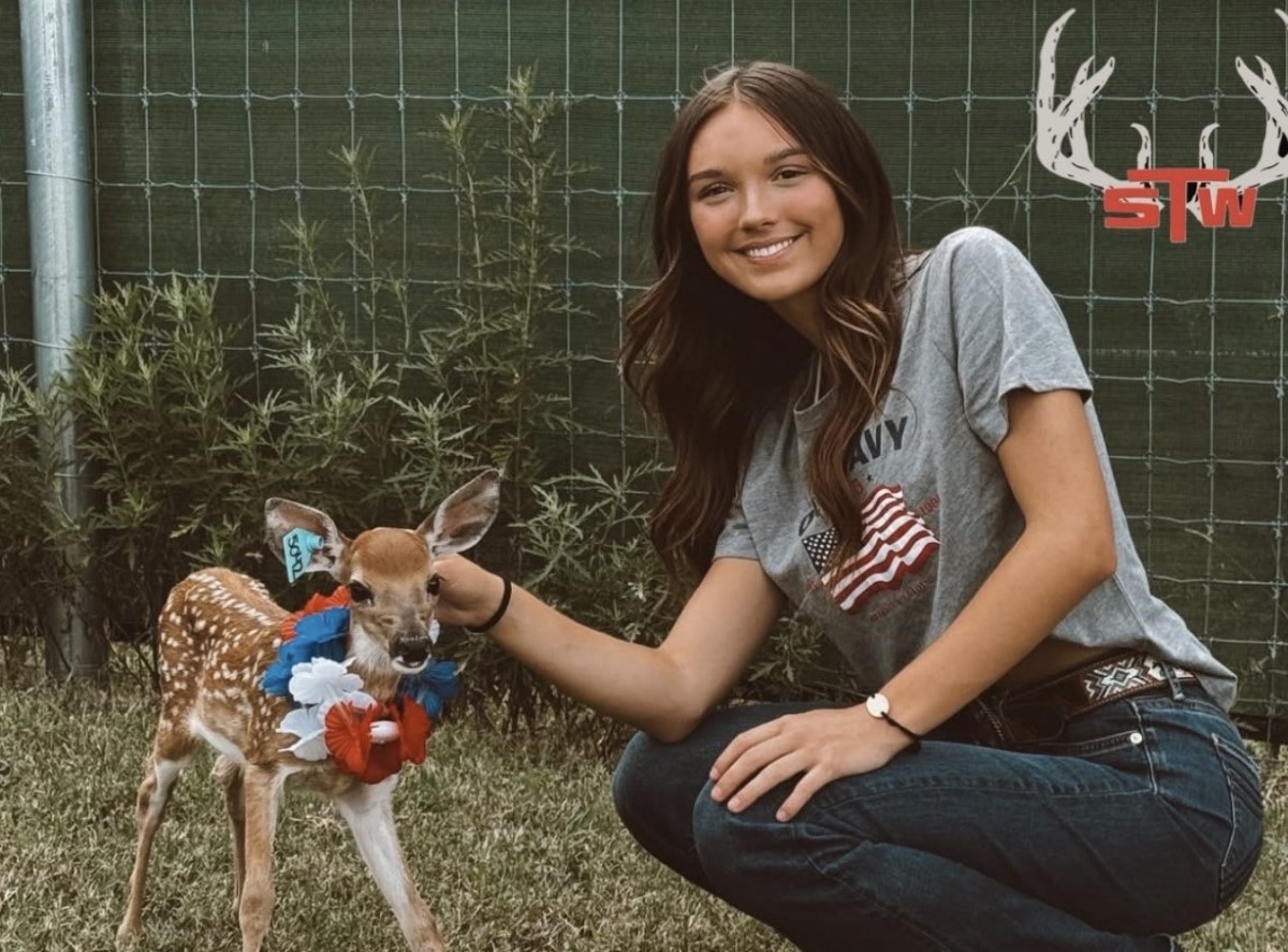 Young woman crouching and smiling while petting a small fawn wearing a red, white, and blue floral collar outdoors by a green fence.