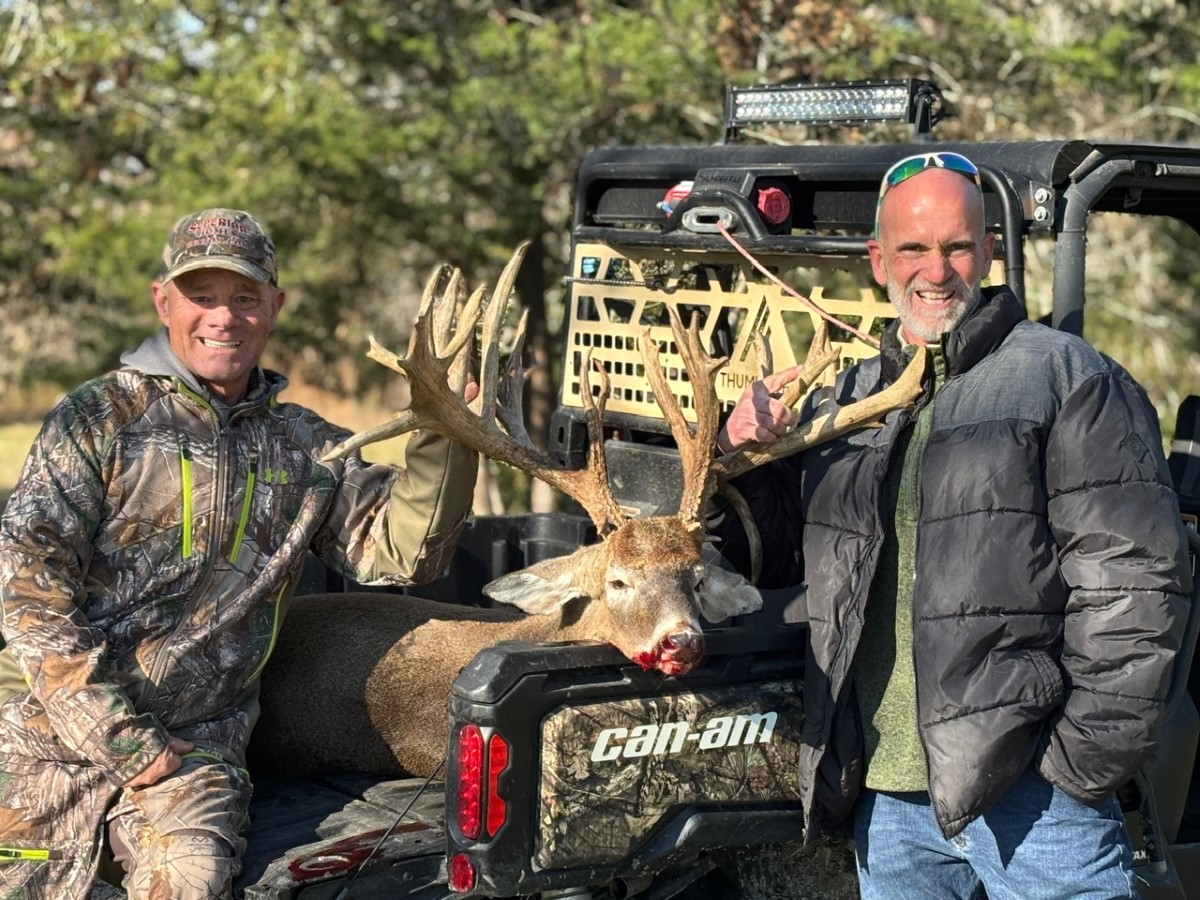 Two men smiling beside a large deer with impressive antlers on a Can-Am vehicle in an outdoor wooded setting.