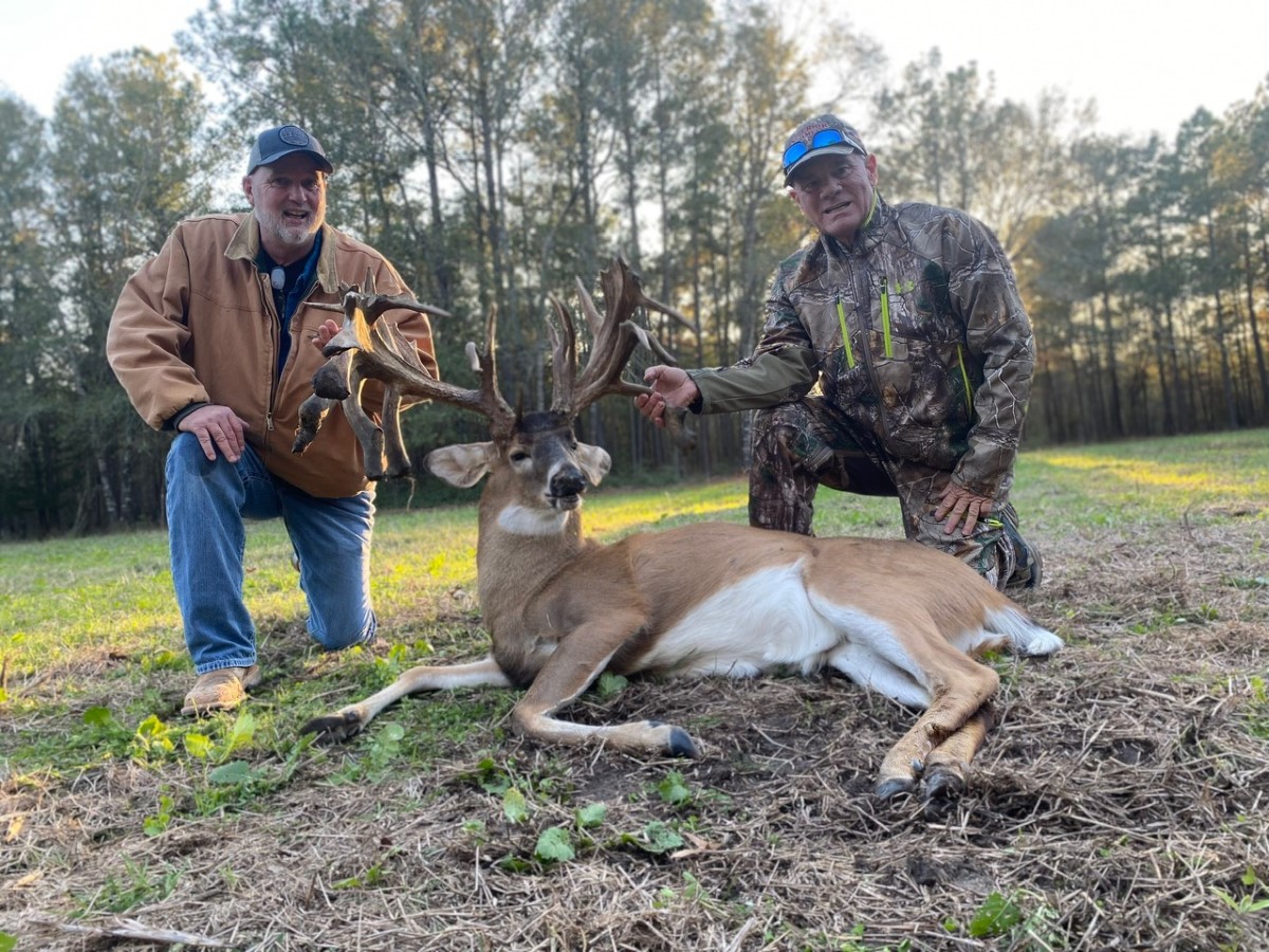 Two men kneeling beside a large buck deer with impressive antlers in an open field near trees.