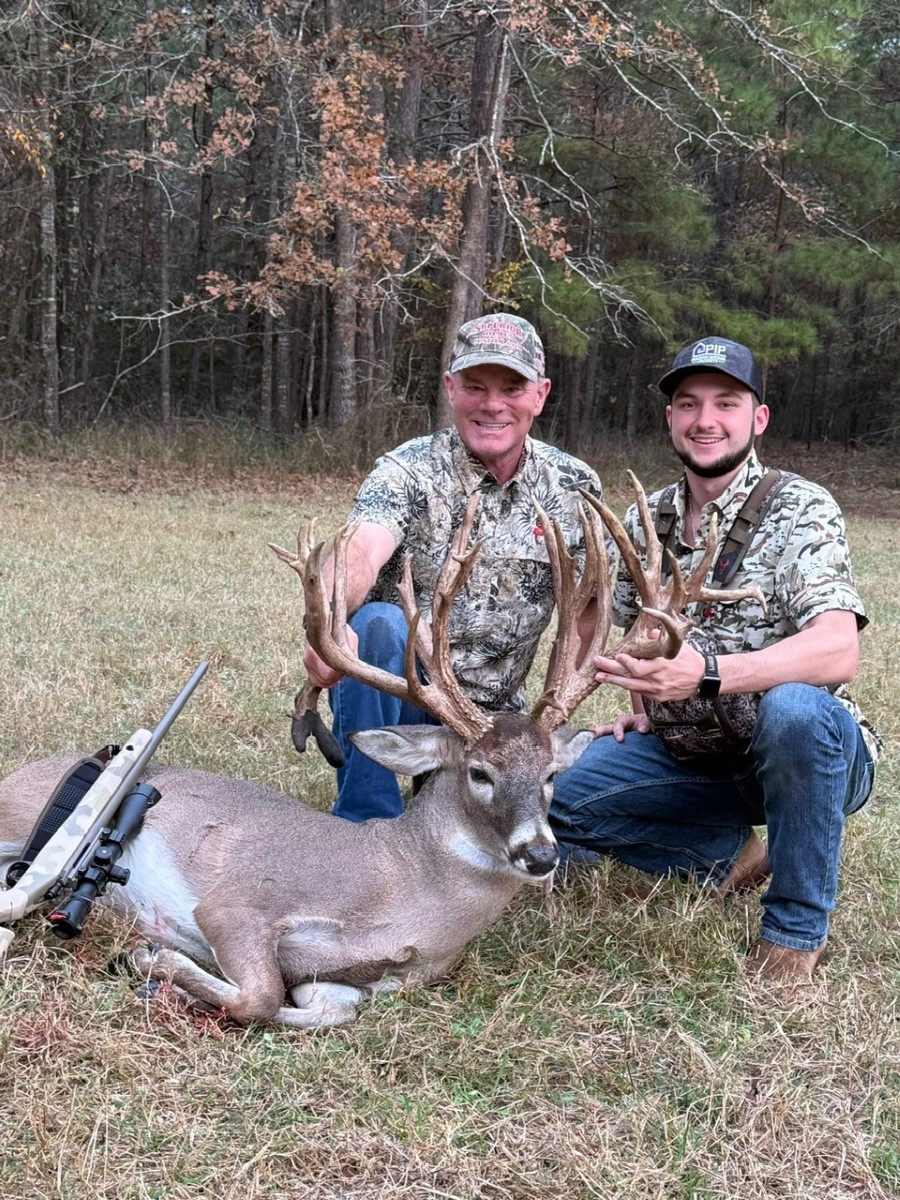 Two hunters kneeling behind a large buck with impressive antlers, lying on grass with a rifle resting beside it.