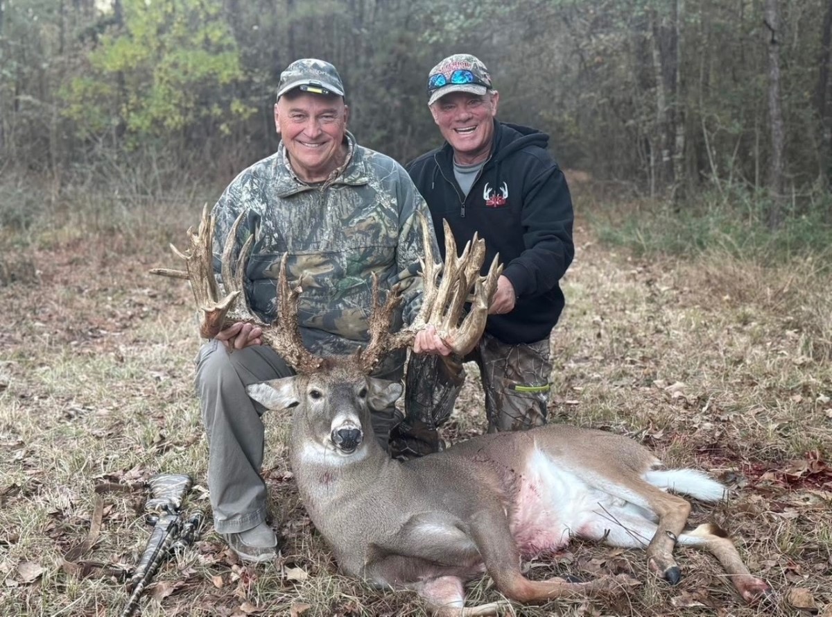 Two men in camouflage and outdoor clothing posing with a large buck deer with massive antlers on the ground.