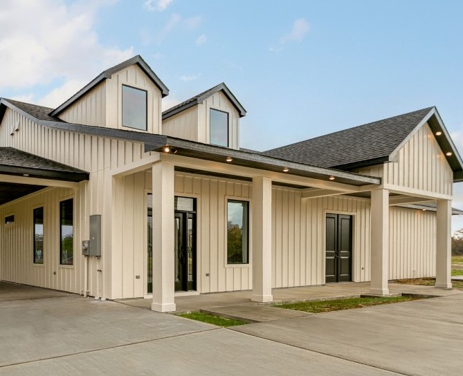 Modern cream-colored barn with vertical siding, dormer windows, covered porch, and concrete driveway under a partly cloudy sky.