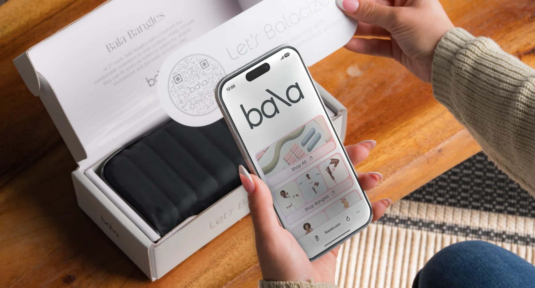 Person holding a phone showing the Bala fitness FlowHub with a box of Bala Bangles in the background and an insert with a Flowcode.