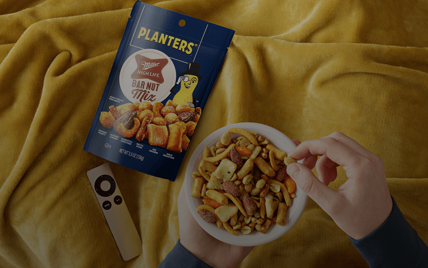 Hands holding a bowl of Planters Miller High Life Bar Nut Mix with an open snack bag and a remote on a yellow blanket.