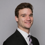 Smiling young man in a black suit with a light purple striped tie against a gray background.