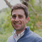 Smiling man with short brown hair wearing a gray quarter-zip sweater outdoors with greenery background.