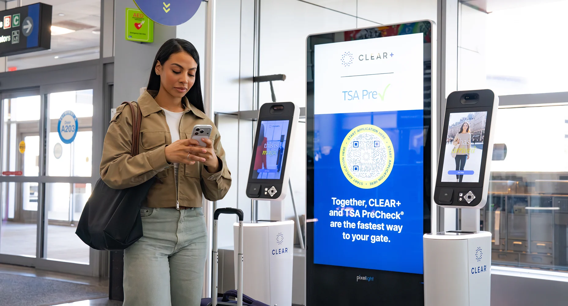 Woman using her phone next to CLEAR+ TSA PreCheck kiosks and sign with Flowcode at airport gate A203.