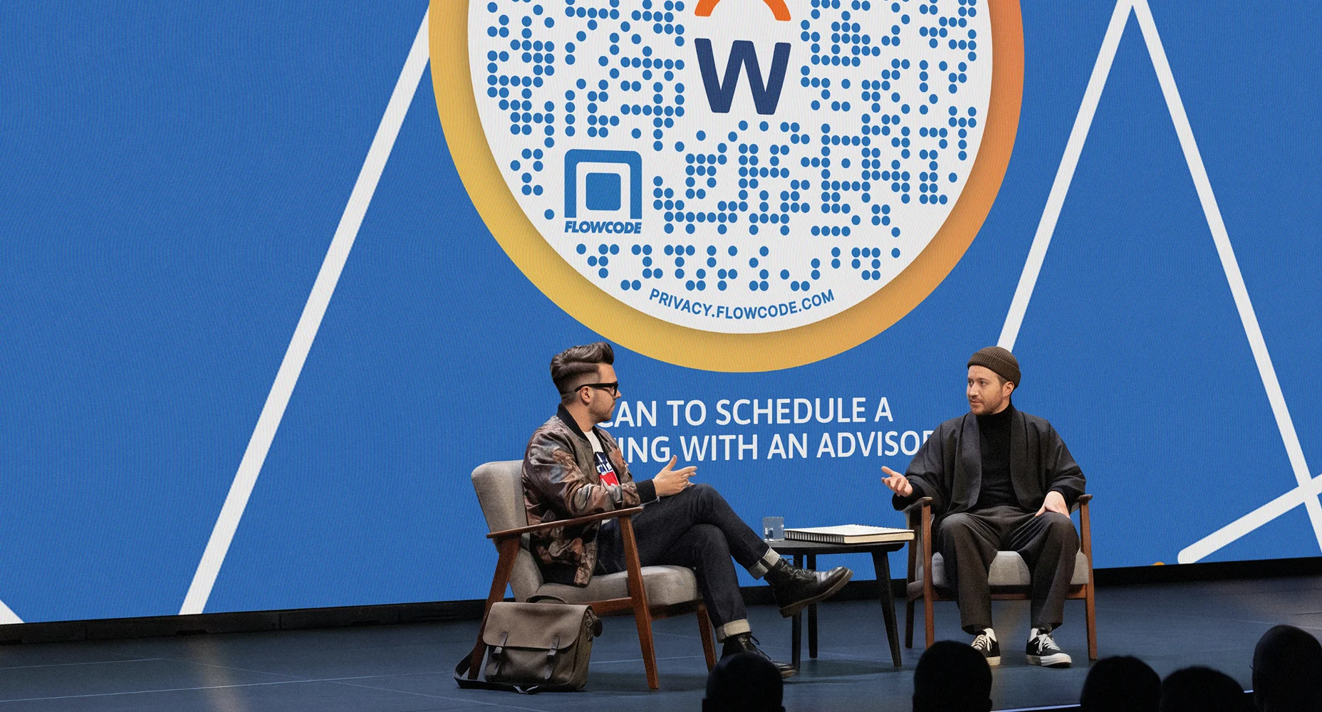 Two men seated on stage having a discussion in front of a large screen displaying a Flowcode.