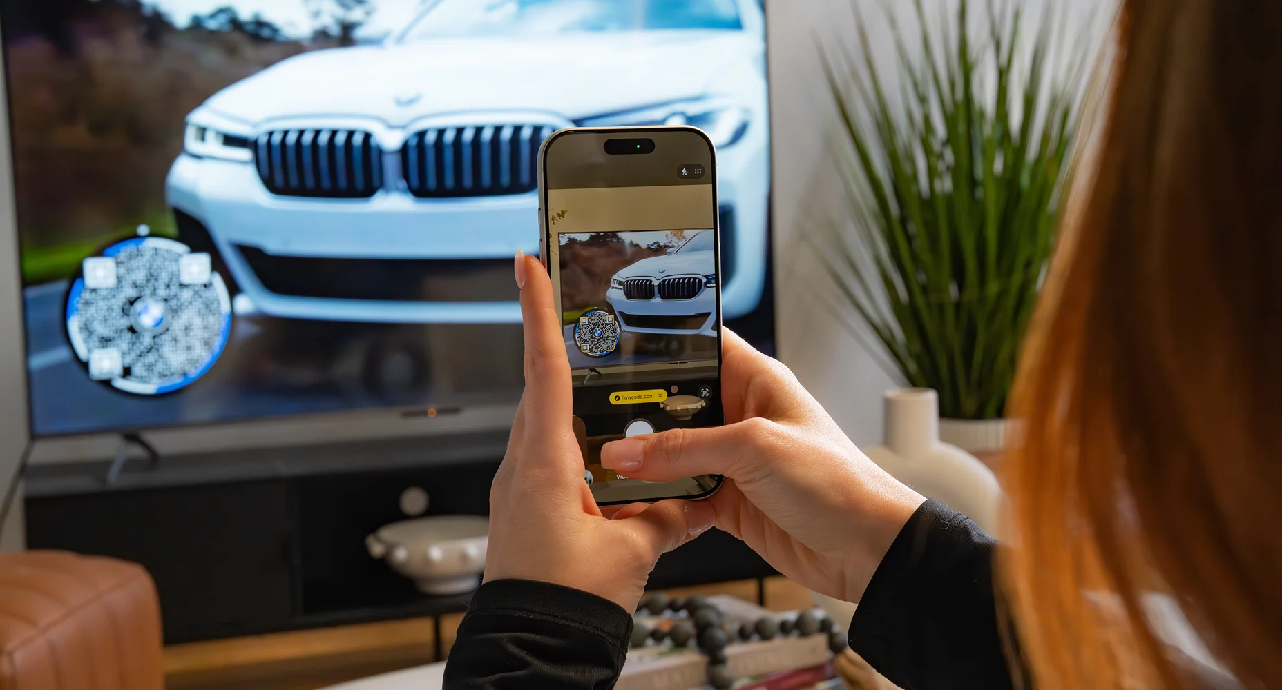 Person using a smartphone to scan a Flowcode on a TV screen showing a white BMW car front.