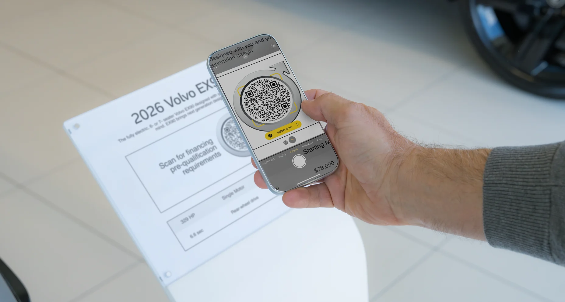 Hand holding a smartphone scanning a Flowcode on a 2026 Volvo EX90 information sheet.