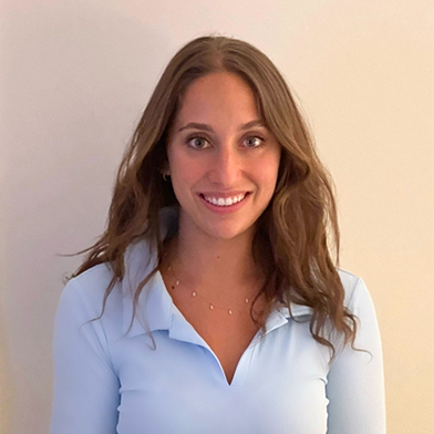 Smiling young woman with long brown hair wearing a light blue top against a beige background.