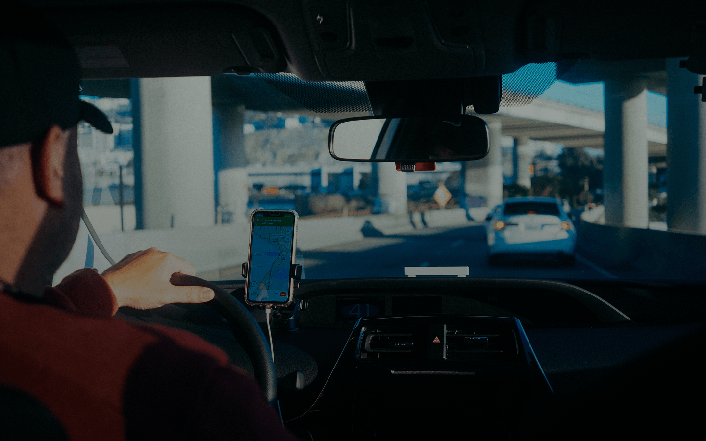 Driver holding steering wheel inside car with phone mounted showing navigation map ahead.