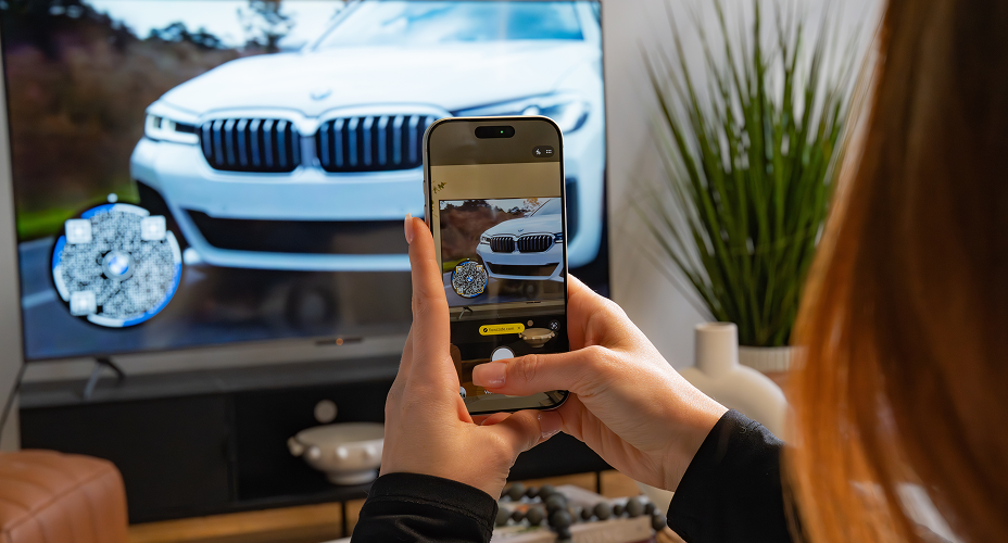 Person holding smartphone scanning Flowcode displayed on TV screen showing front of white BMW car.