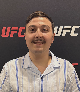 Man with mustache smiling in front of a black UFC backdrop with red and white UFC logos.
