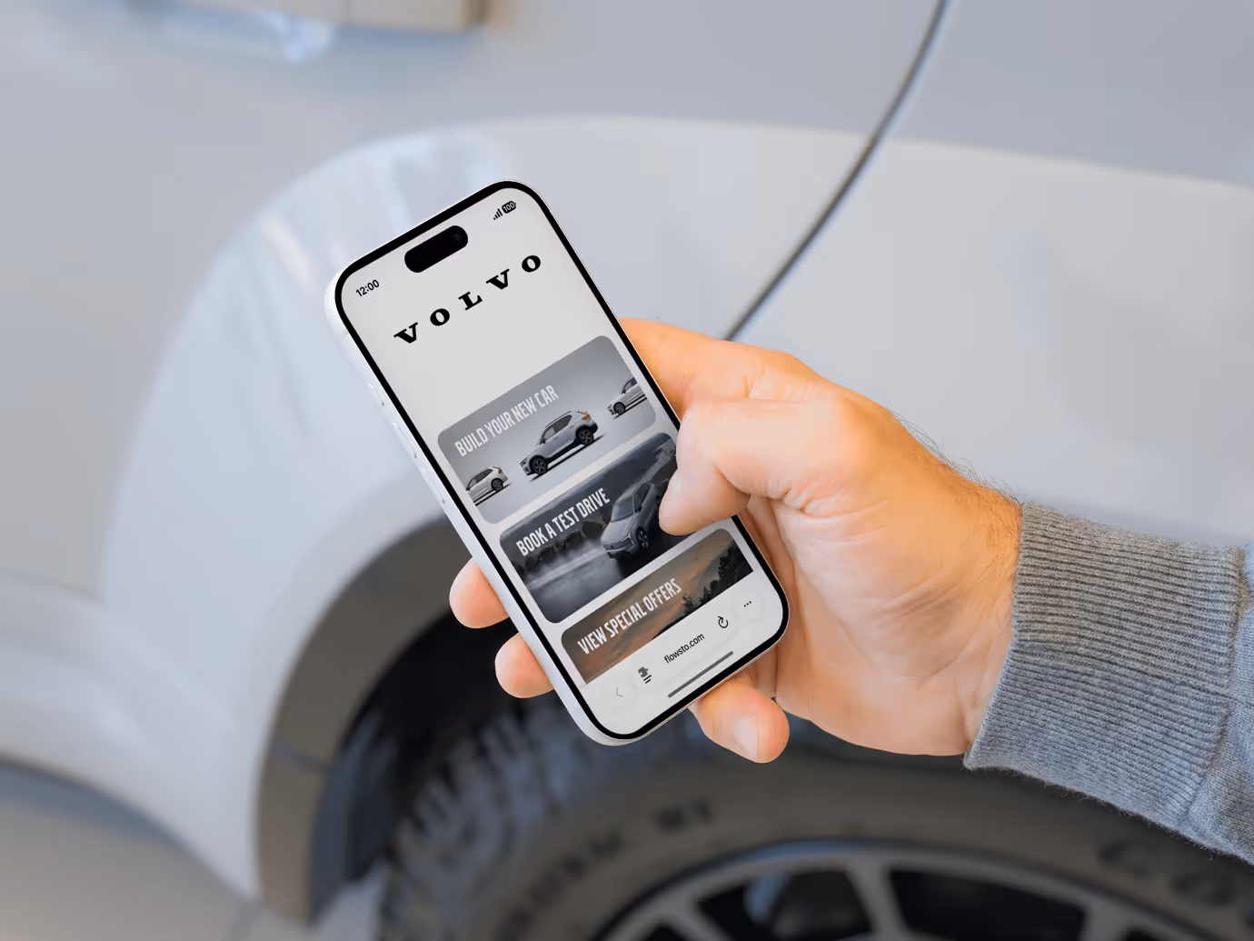 A person holding a smartphone showing a Volvo FlowHub with options to build a car, book a test drive, and view special offers.