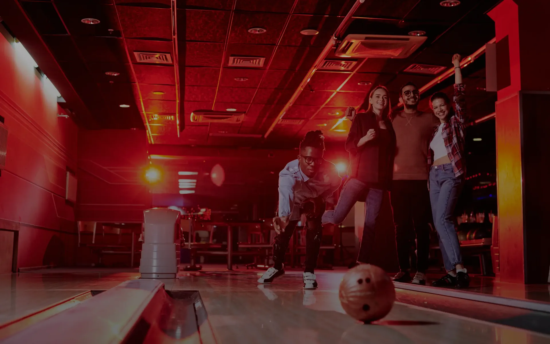 Group of friends bowling indoors with one person rolling the ball and others cheering.