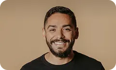 Smiling man with short dark hair and beard wearing a black t-shirt against a warm beige background.