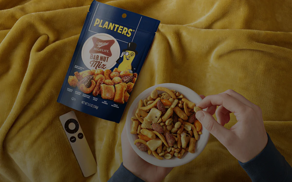 Hands holding a bowl of Planters Miller High Life Bar Nut Mix with an open snack bag and a remote on a yellow blanket.