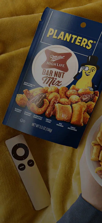 Hands holding a bowl of Planters Miller High Life Bar Nut Mix with an open snack bag and a remote on a yellow blanket.