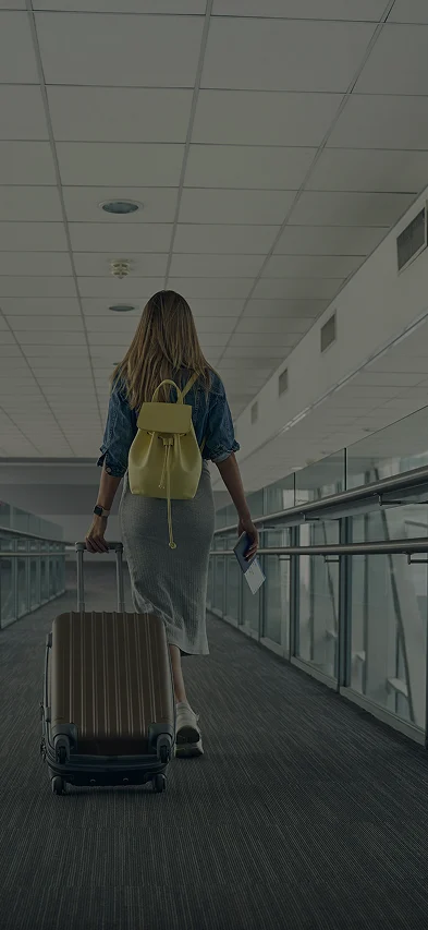 A woman with a yellow backpack walks while rolling carry on luggage behind her down an airport corridor.
