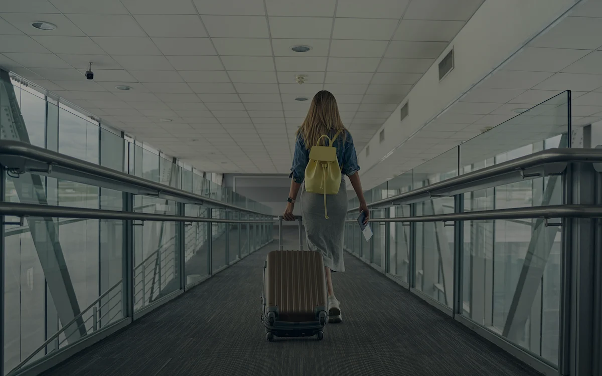 A woman with a yellow backpack walks while rolling carry on luggage behind her down an airport corridor.
