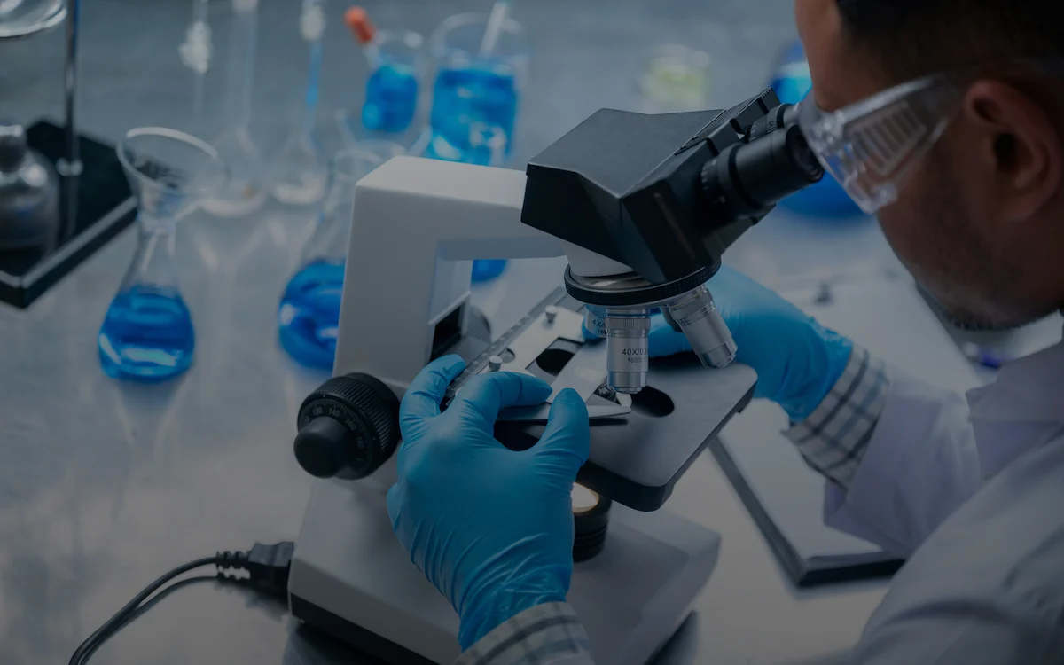 Scientist in blue gloves and safety goggles examining a sample under a microscope in a laboratory setting with blue liquid reagents in the background.