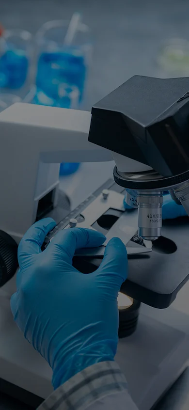 Scientist in blue gloves and safety goggles examining a sample under a microscope in a laboratory setting with blue liquid reagents in the background.