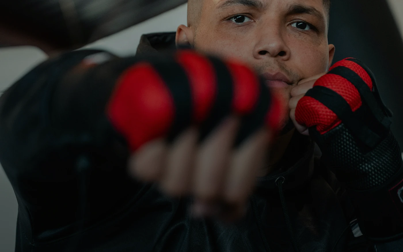 A boxer with a buzz cut and red boxing gloves on holds his hands below his eyes in front of his face. 