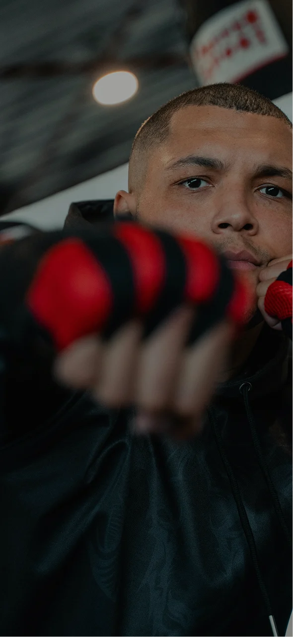 A boxer with a buzz cut and red boxing gloves on holds his hands below his eyes in front of his face. 