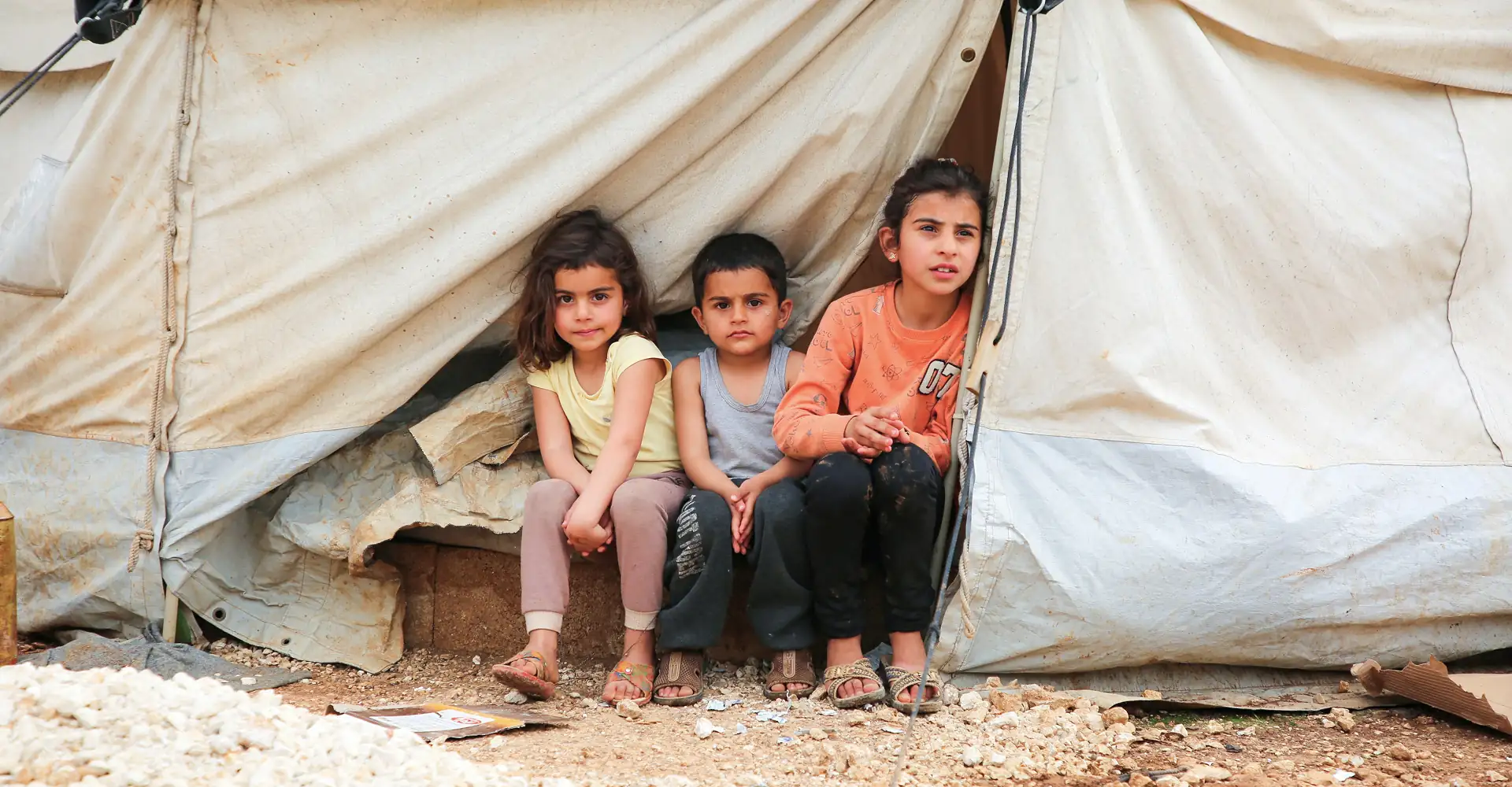 Three children affected by war sitting together inside a tent