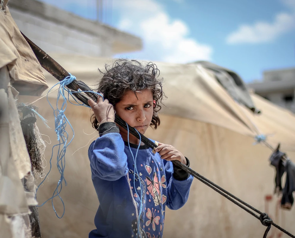 Child living in a refugee camp standing near tents holding a wooden pole