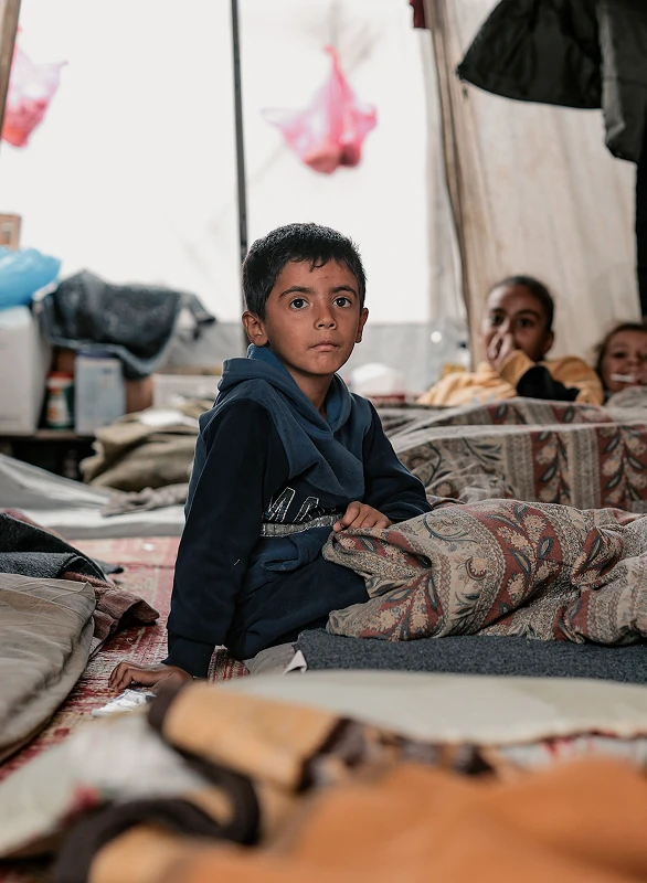 Child sitting on a bed inside a tent with others nearby