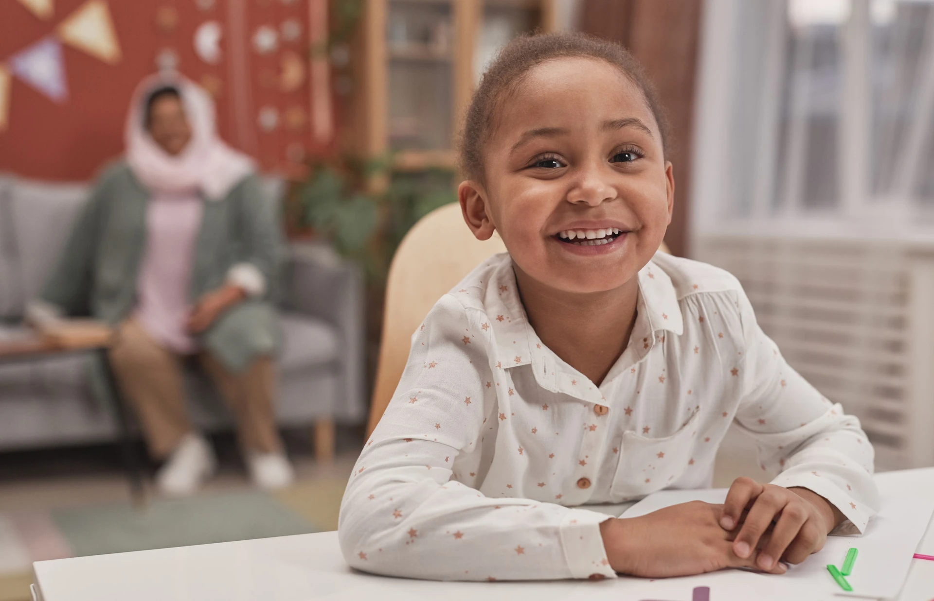Smiling child sitting at a table with an adult in the background