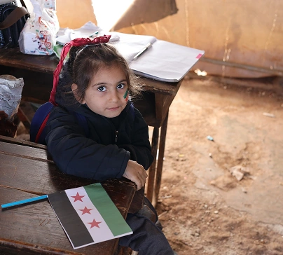 Young girl sitting at a wooden school desk in a classroom, wearing a winter coat, with a small flag and papers on the desk
