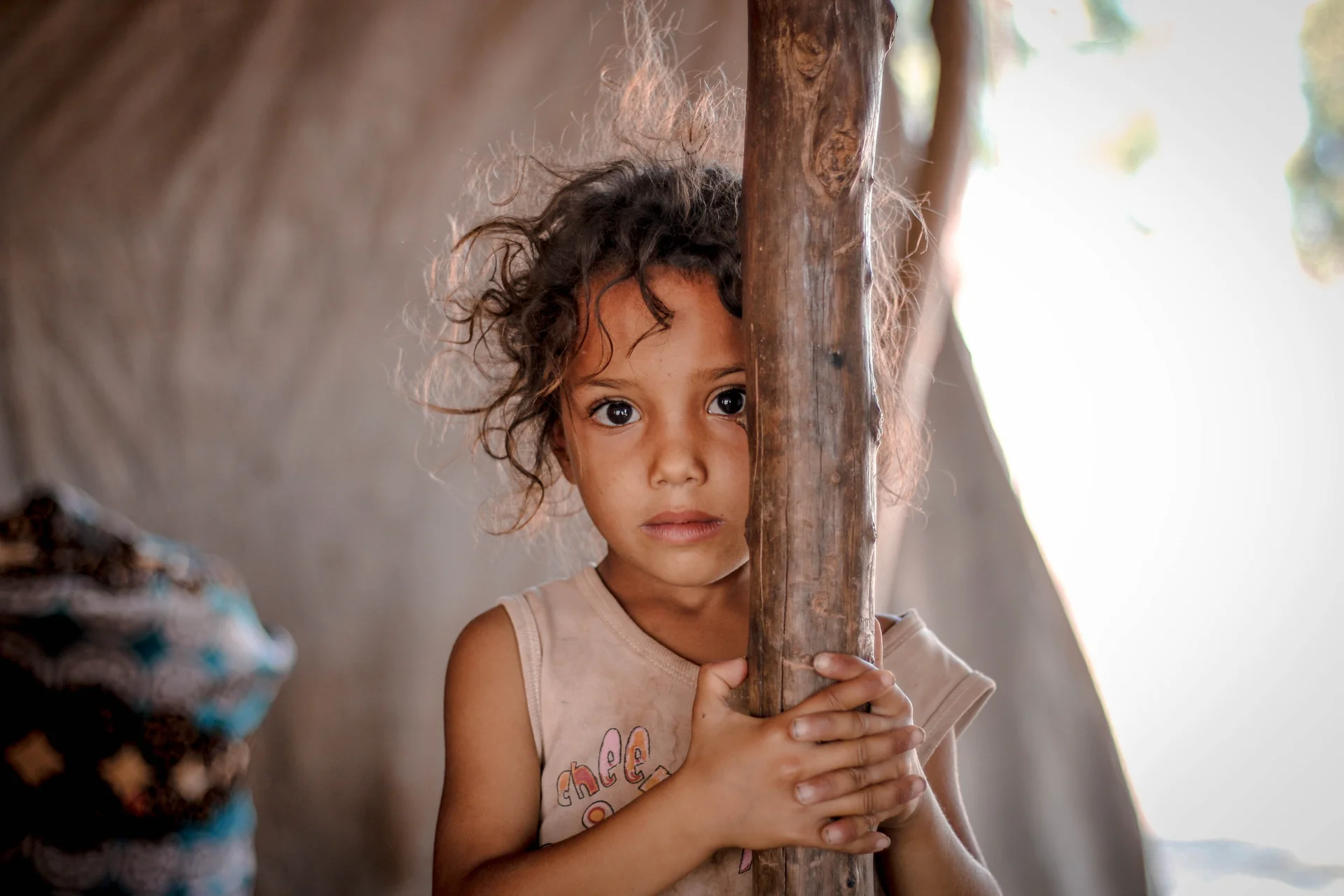 Young girl with curly hair holding a wooden pole inside a tent, looking directly at the camera
