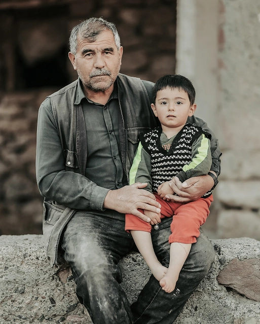 Elderly man sitting outdoors with a young boy on his lap, both looking at the camera