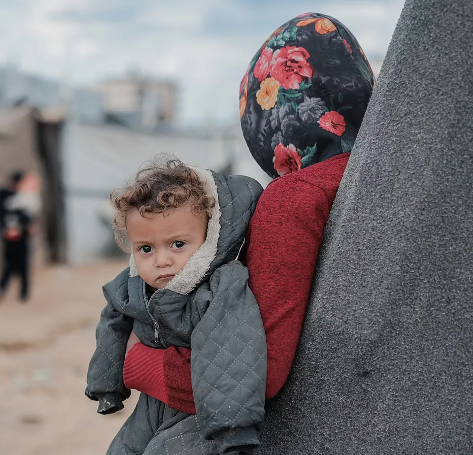 Mother carrying her toddler on her back at a displacement camp, child looking toward the camera