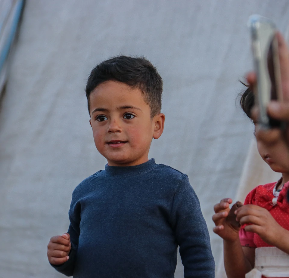 Young boy in a refugee camp setting looking up, standing in front of a tent with another child nearby