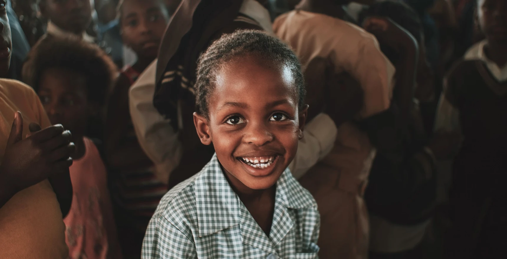 Young child in a checkered school uniform smiling at the camera, surrounded by other children indoors