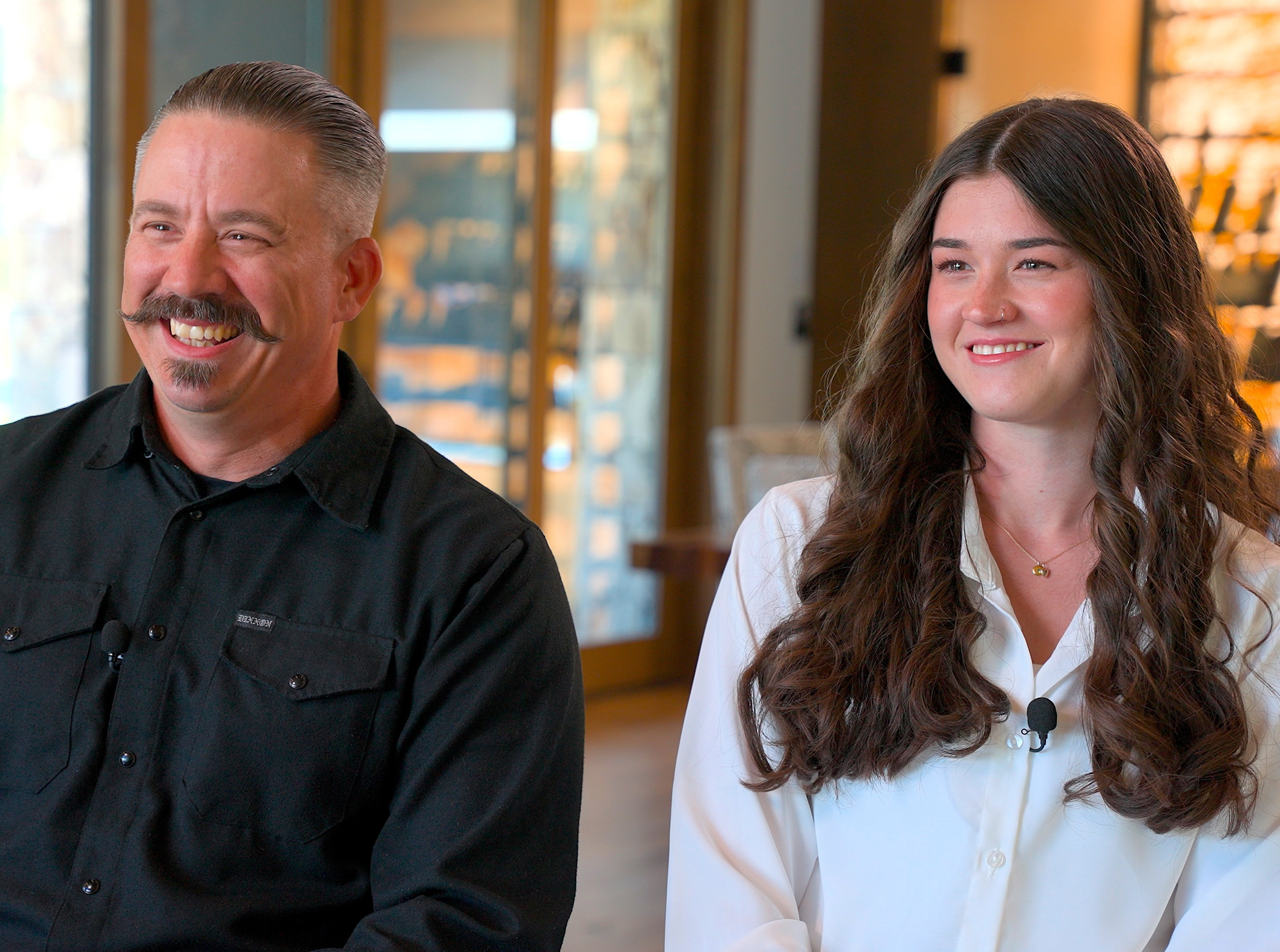 A smiling man with a mustache in a black shirt and a woman with long brown hair in a white blouse.