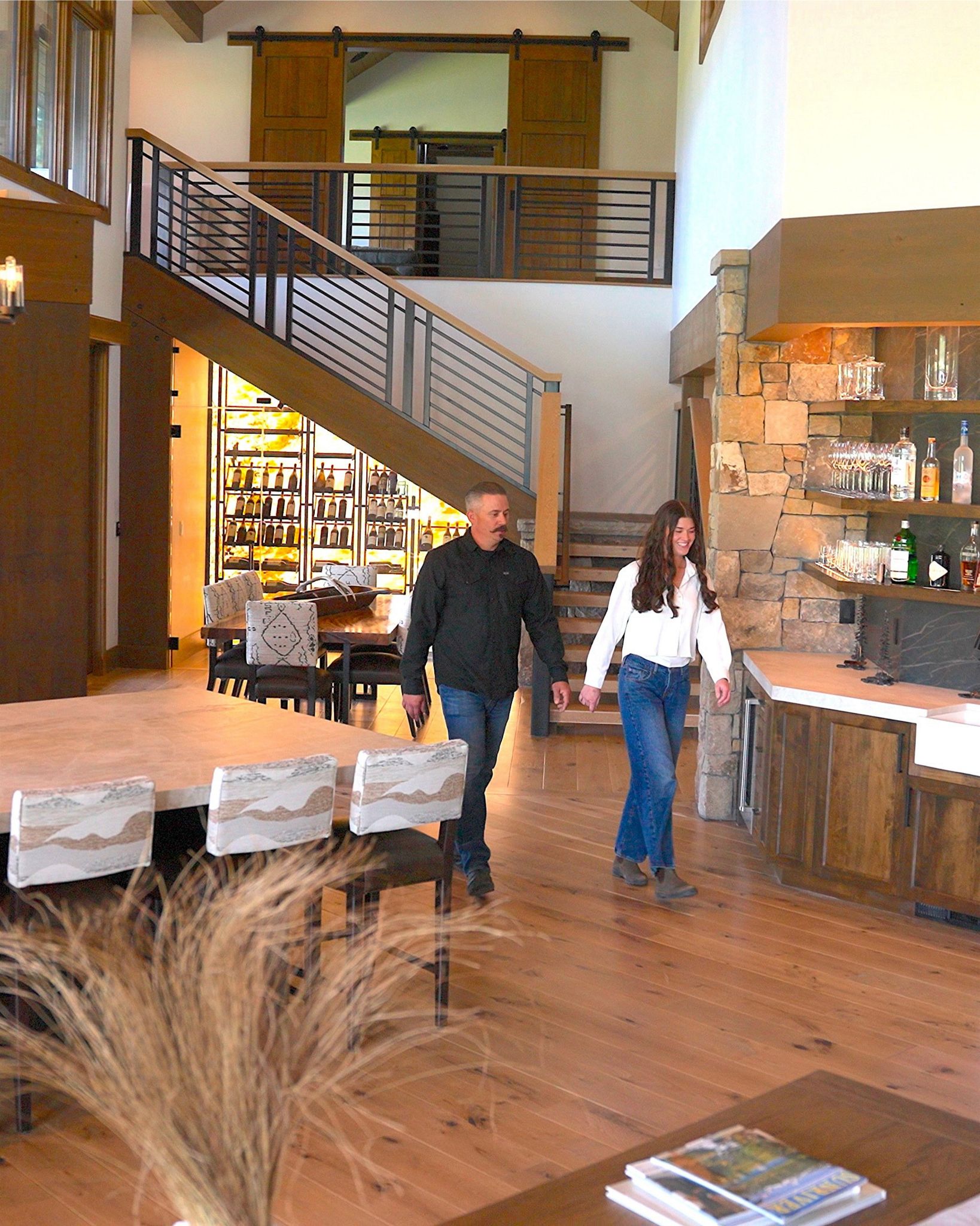 Man and woman walking hand in hand inside a modern home with wooden floors and wine storage.
