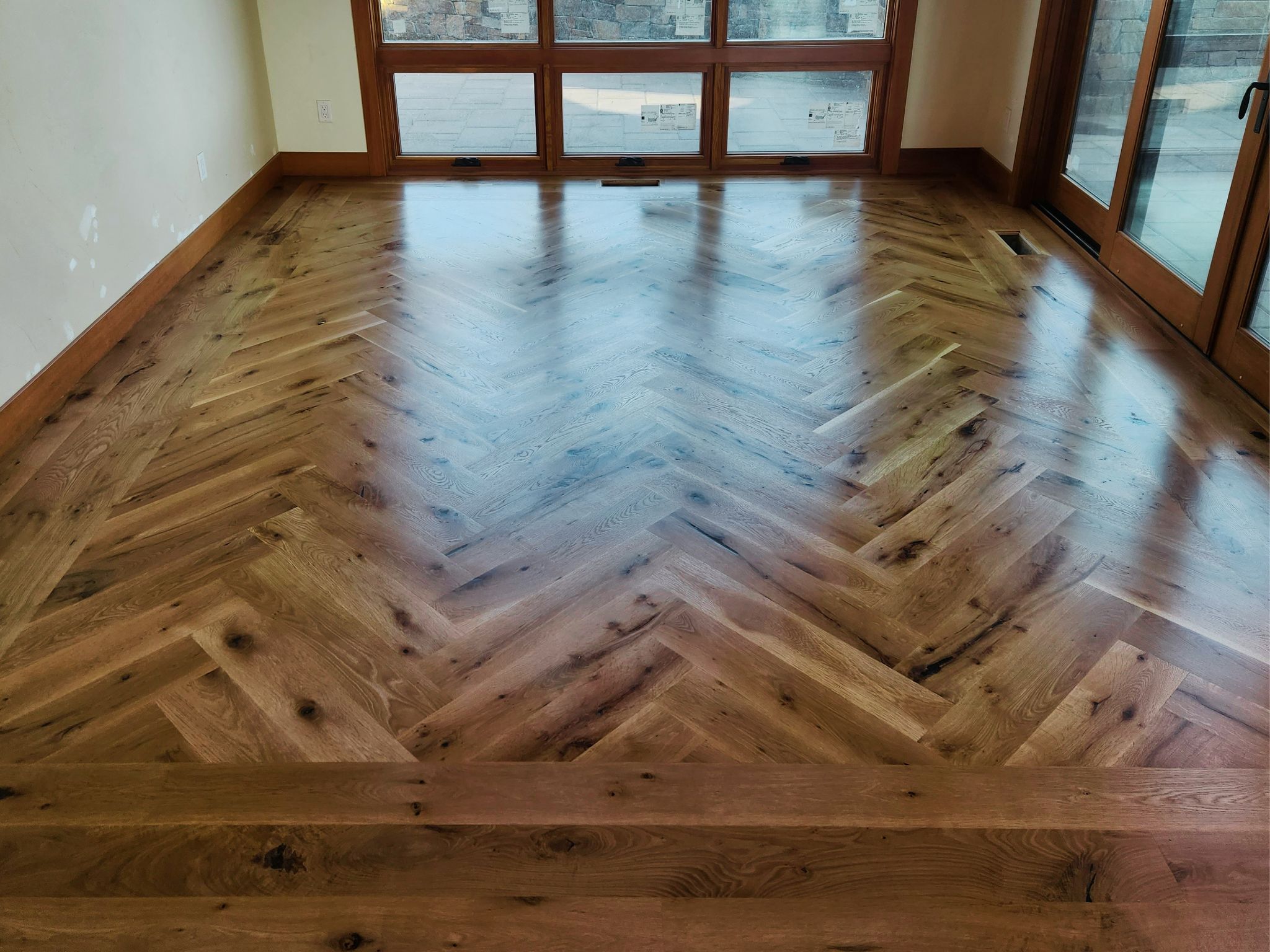 Polished wood floor with herringbone pattern in a room with large windows and glass doors.