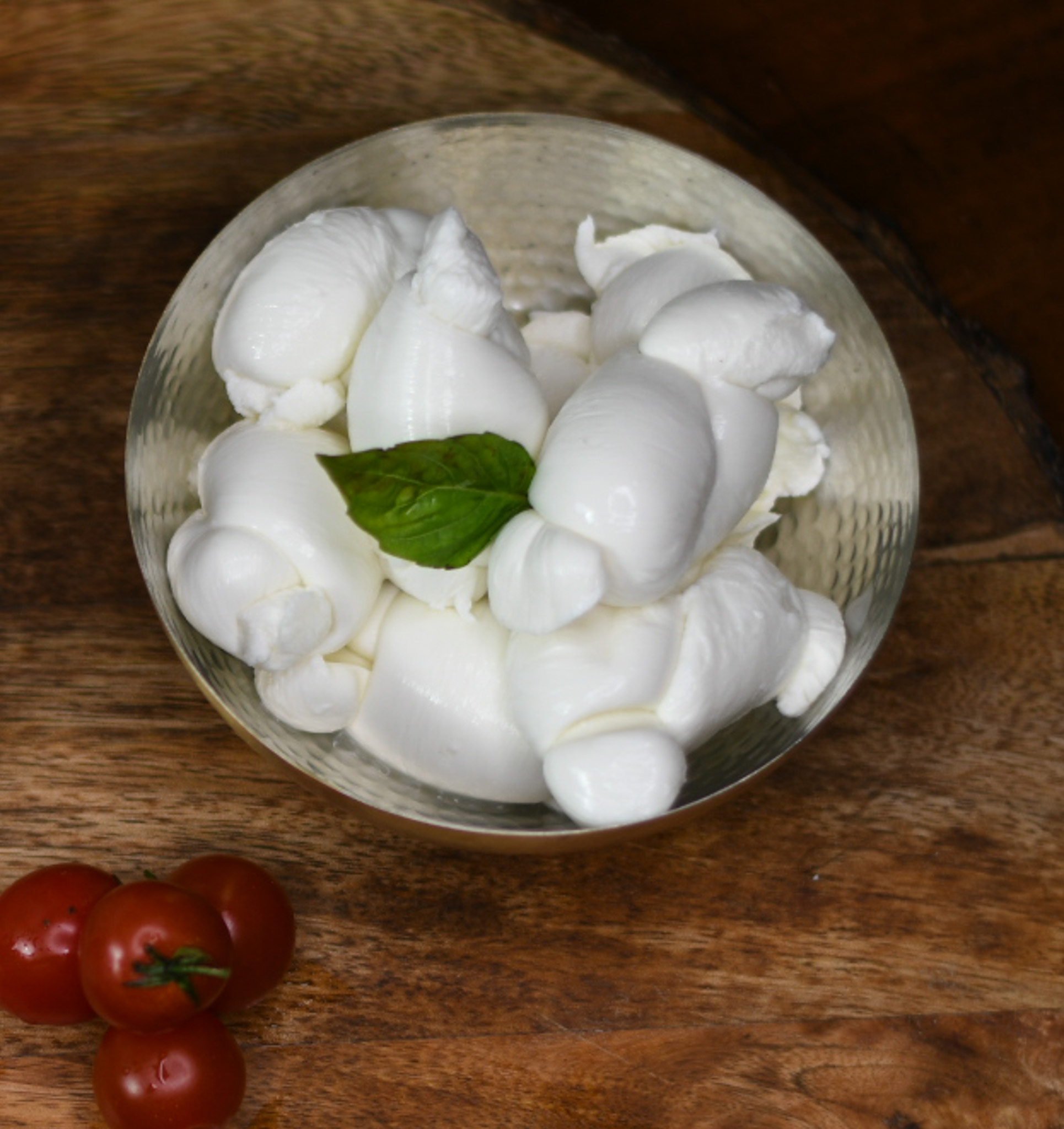 Glass bowl of fresh mozzarella balls garnished with a basil leaf, next to cherry tomatoes on wood.