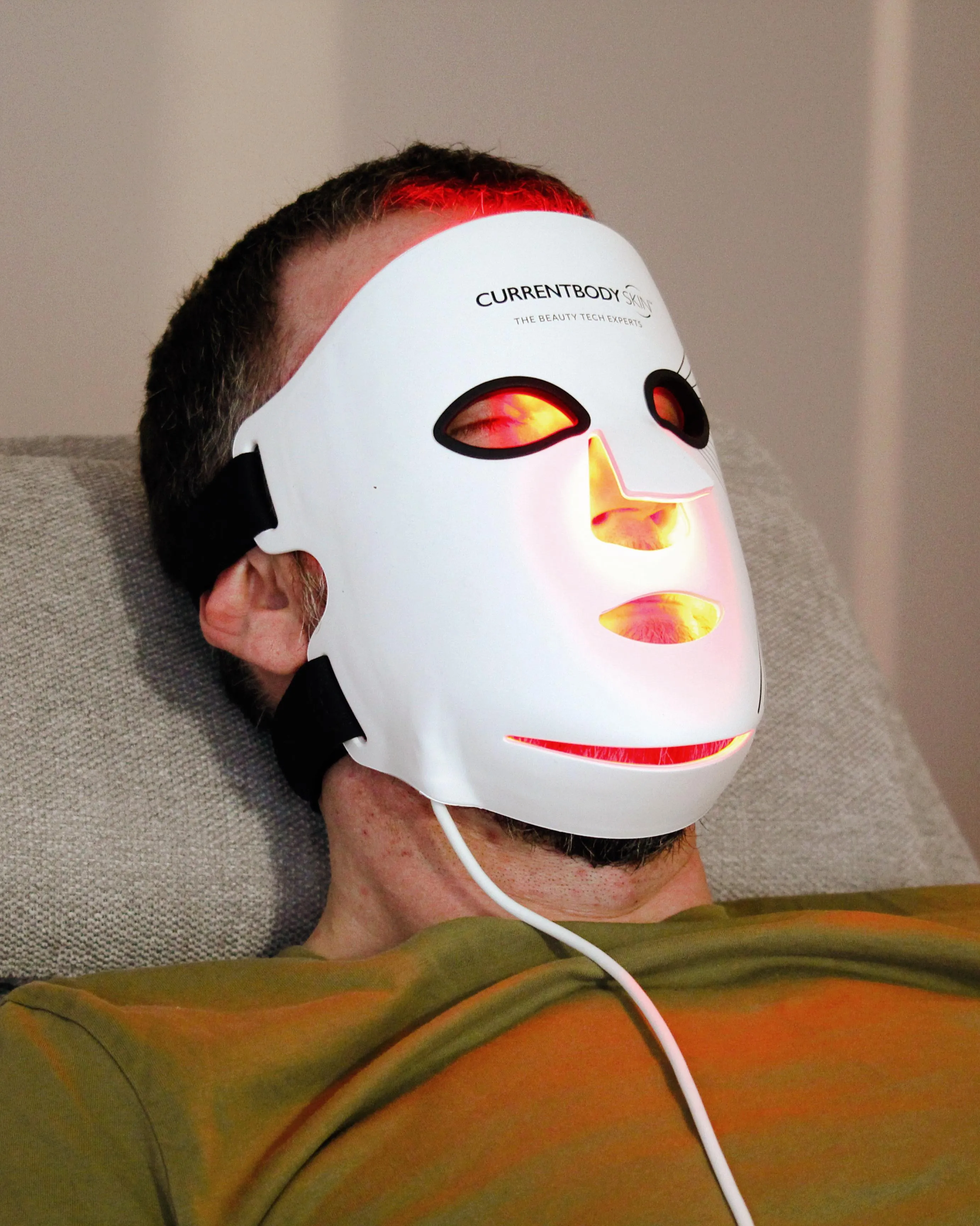 Ein Mann liegt zurückgelehnt da und trägt eine weiße LED-Lichttherapiemaske mit der Aufschrift „CurrentBody Skin“.