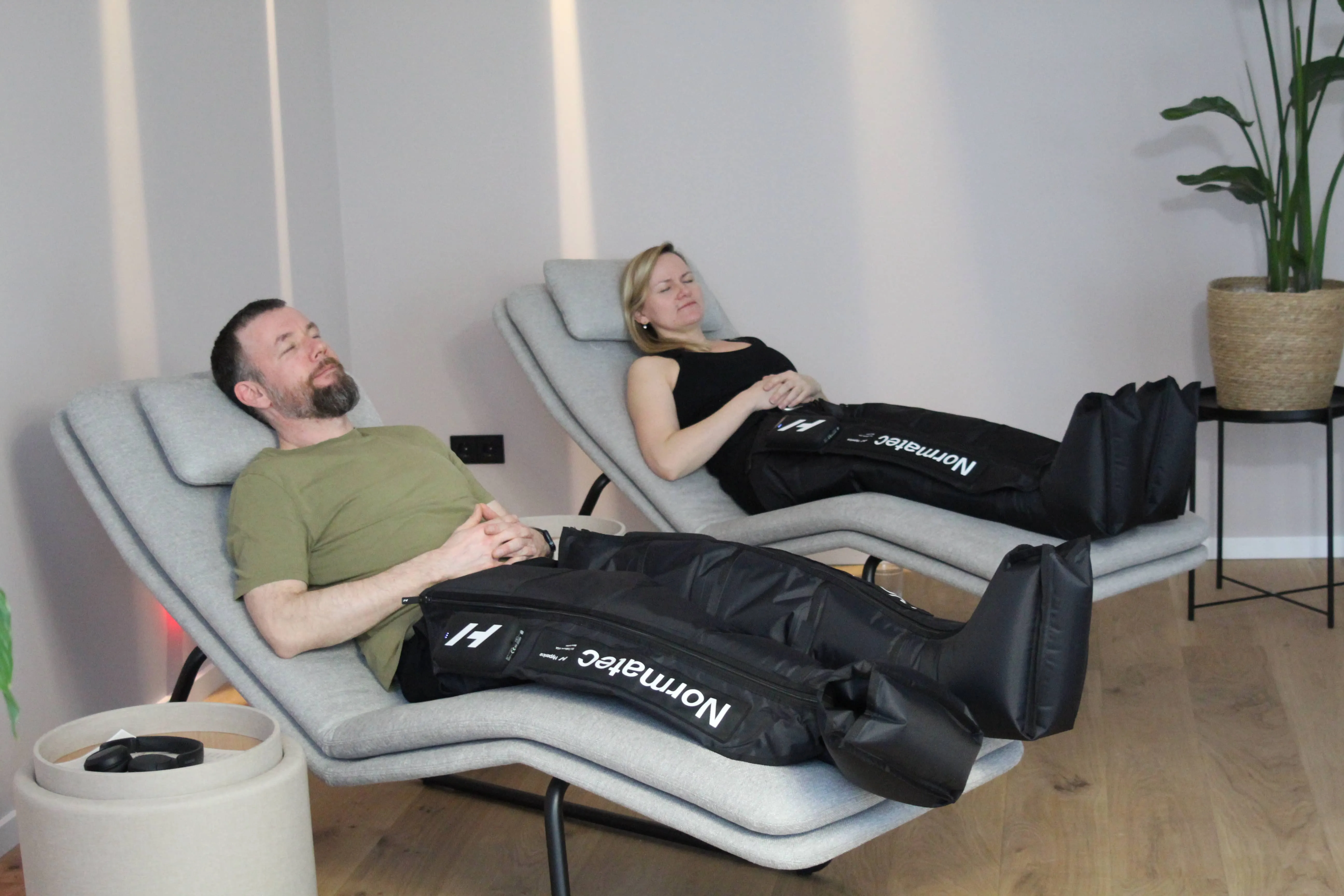 Man and woman relaxing on chairs wearing Normatec compression leg sleeves in a modern room.
